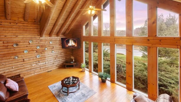 Interior de una cabaña de troncos con chimenea, ventanas amplias, pisos de madera y ventiladores de techo.