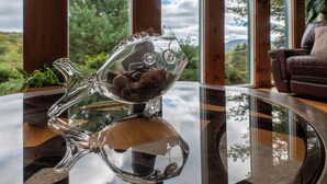 Una escultura de vidrio con forma de pez sobre una mesa de vidrio reflectante, con vistas a la vegetación y montañas a través de amplios ventanales.