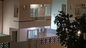 Un edificio residencial de varios pisos con balcones, una entrada protegida por una reja y un árbol en primer plano.