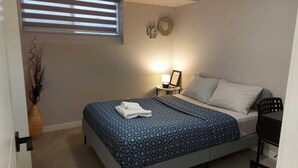 Een slaapkamer met een bed, nachtkastje, lamp, decoratieve spiegel en een raam met zonwering.