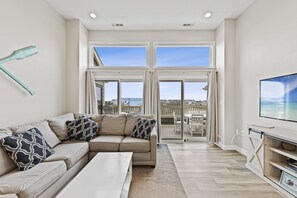 A modern living room with a large sofa, a flat-screen TV, and a balcony with a view of the sea. - Gone Coastal Top Floor, Bay Views, Pool, 120th Street, Quiet Neighborhood (Ocean City)