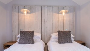 A room with two beds, each with white bedding and a knitted throw pillow. There are wooden nightstands with lamps on either side of the beds. - Sailmaker's Cottage (Lymington)