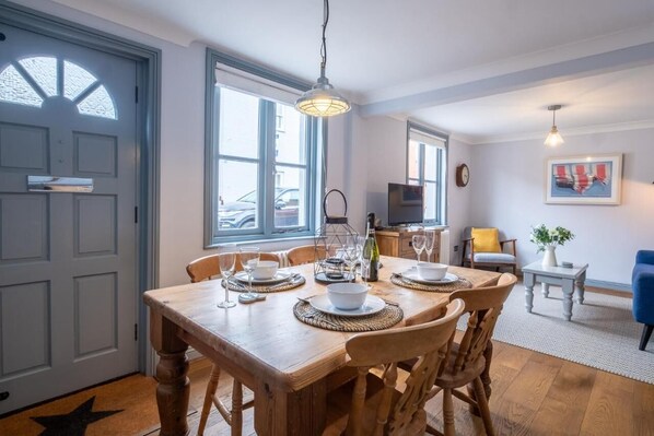 A dining area with a wooden table set for four, surrounded by chairs. There is a kitchen area with a refrigerator and a microwave in the background.