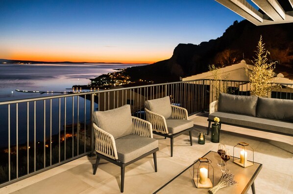 A balcony with wicker chairs and a sofa, a table with candles and glasses, and a view of the sea and city lights at dusk.