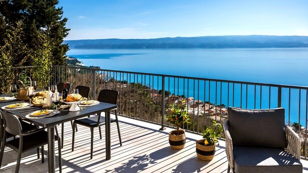 Une terrasse avec une table dressée pour le dîner, offrant une vue sur une étendue d’eau et sur des collines lointaines.