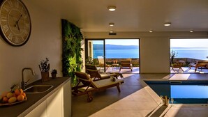 Une cuisine moderne avec une grande horloge, un coin repas offrant une vue sur la mer et un espace piscine avec des chaises longues.