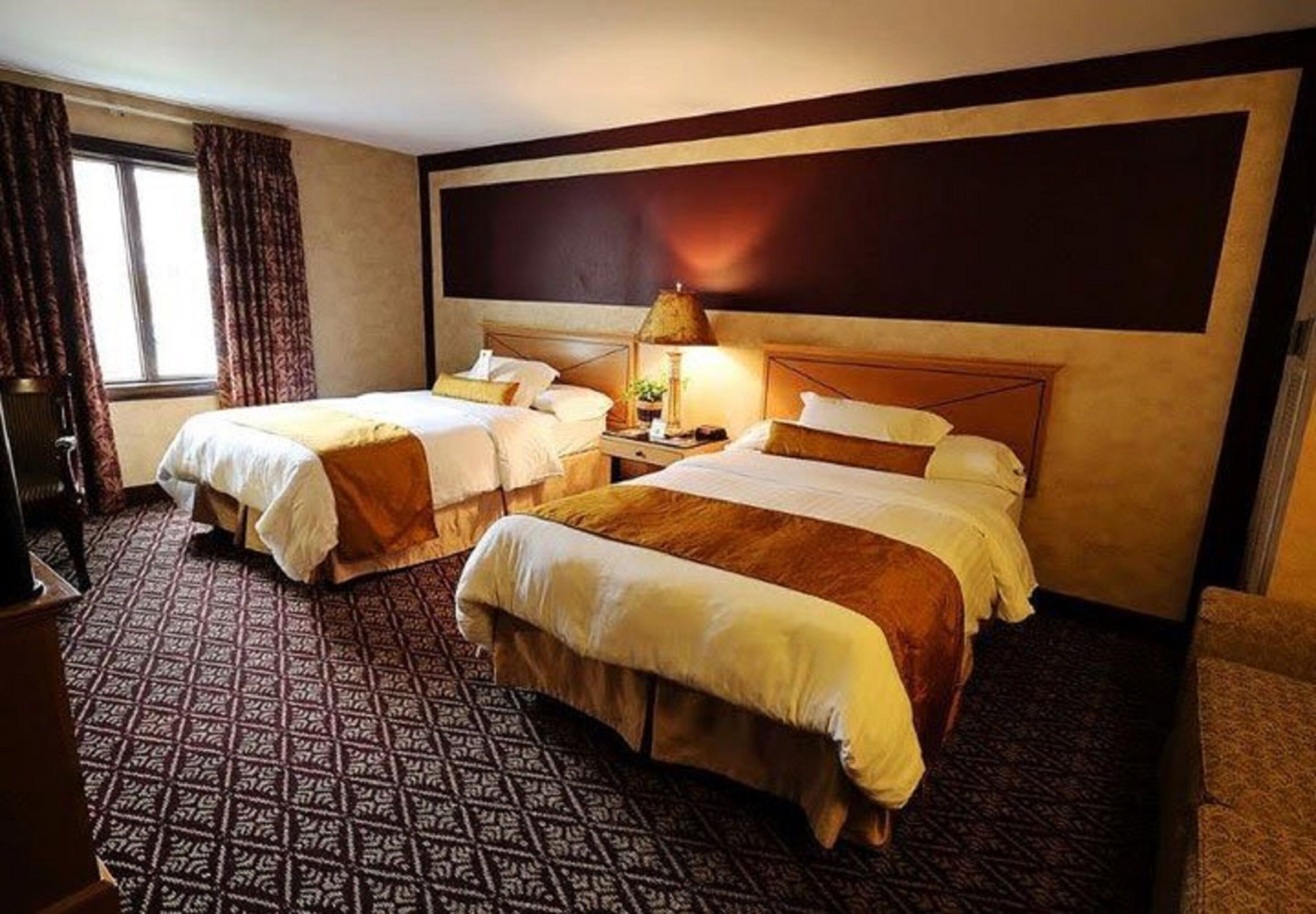 A hotel room with two beds, a nightstand with a lamp, patterned carpet, and a large dark wall.