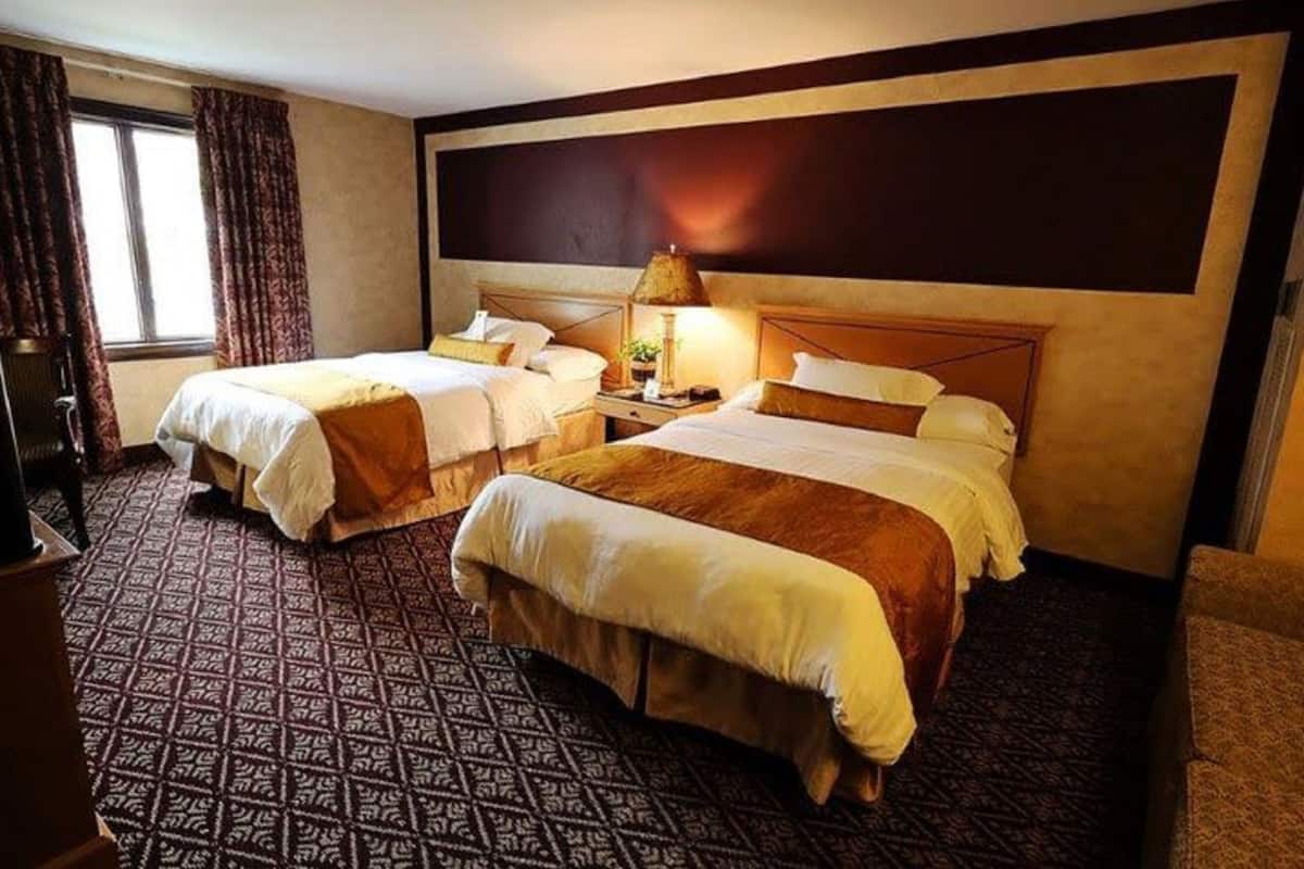 A hotel room with two beds, a nightstand with a lamp, patterned carpet, and a large dark wall.