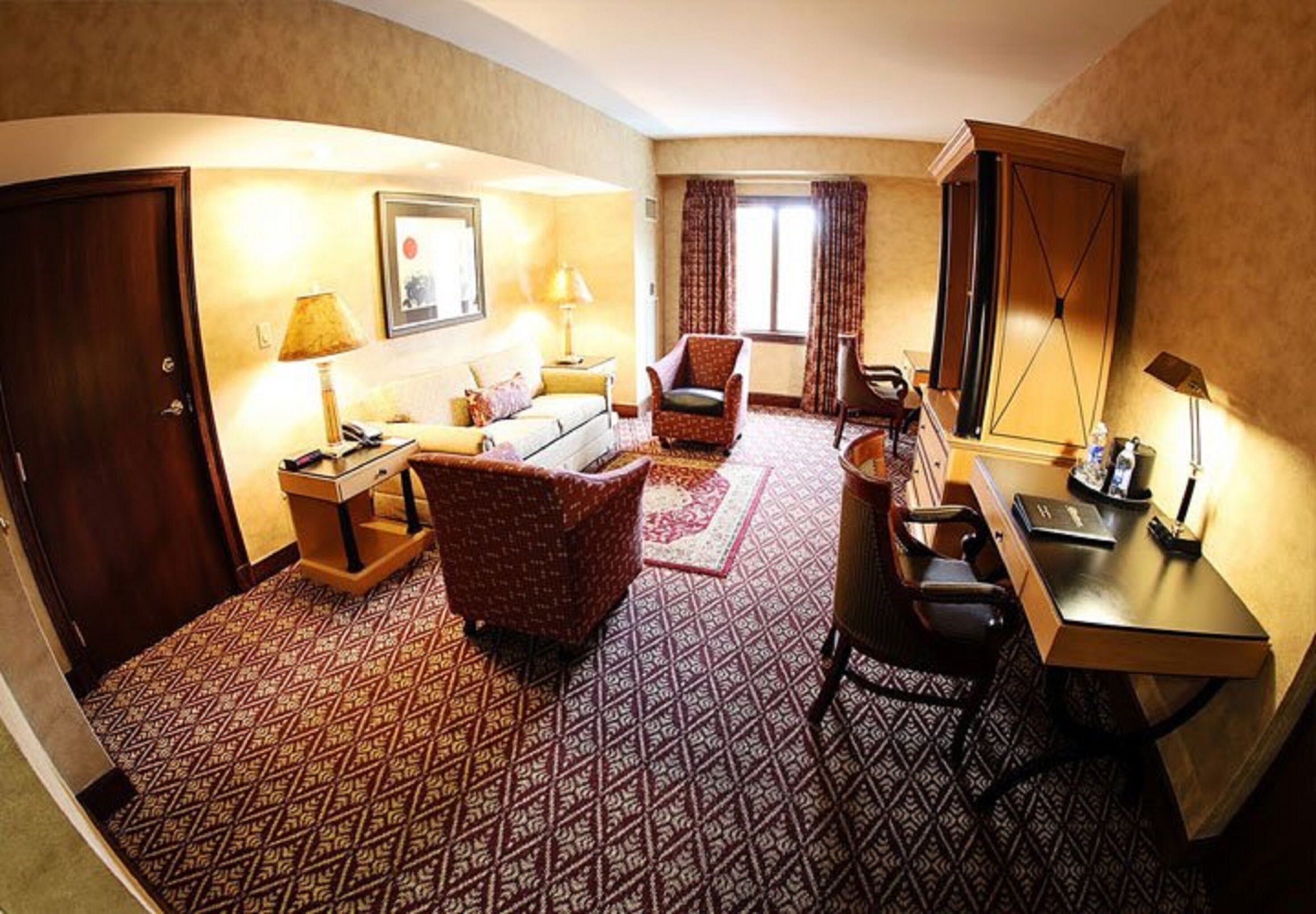 A hotel room with a patterned carpet, a sofa, a chair, a desk, a lamp, a mirror, and a door.