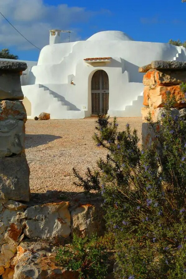 A white building with a central arched doorway, surrounded by stone walls and gravel ground.