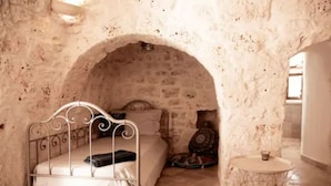 Una habitación con pared de piedra, una cama de metal, una mesa redonda y un pequeño arco.