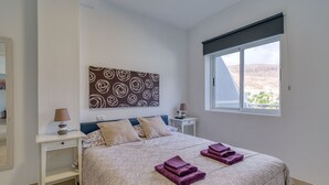 A bedroom with a large bed, a nightstand, a lamp, a wall art piece, and a window with a view of the landscape. - 20 meters from the beach, private parking and AC (Mogán)