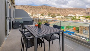 A balcony with a table and chairs, overlooking a pool and a town with hills in the background. - 20 meters from the beach, private parking and AC (Mogán)