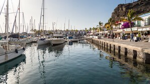 A marina with multiple boats docked, a pier with people and shops, and a rocky cliff in the background. - 20 meters from the beach, private parking and AC (Mogán)
