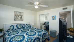 A bedroom with a bed, a ceiling fan, a mirror, a dresser, a desk, and a door leading to another room. - Mescal Loop Hava-Gataway (Lake Havasu City)