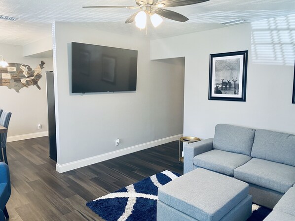 A modern living room with a grey sofa, a flat-screen TV mounted on the wall, a framed picture of deer, and a blue rug with a white cross design. - Beautiful Jacksonville Home (Jacksonville)