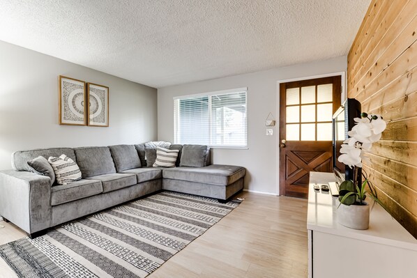 Ein modernes Wohnzimmer mit einem grauen Ecksofa, einem gemusterten Teppich, einer Wand mit Holzpaneelen und einem Fenster mit Jalousien.