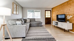 Ein modernes Wohnzimmer mit einem grauen Ecksofa, einer Holz-Akzentwand, einem Flachbildfernseher auf einer weißen Konsole und einem großen Fenster mit Jalousien.