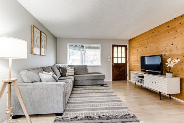 Un salon moderne avec un canapé d’angle gris, un mur d’accent en bois, un téléviseur à écran plat posé sur une console blanche et une grande fenêtre équipée de stores.