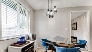 Ein Essbereich mit einem runden Glastisch, blauen Polsterstühlen, einer modernen Leuchte, einem Fenster mit Jalousien und einer Küchenzeile mit Mikrowelle und Wasserkocher.