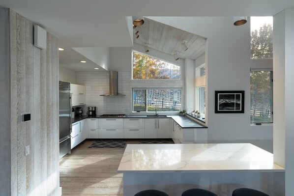A modern kitchen with white cabinetry, a stainless steel refrigerator, and a large window with autumn foliage view. - Stunning Elkhorn Retreat (Sun Valley)