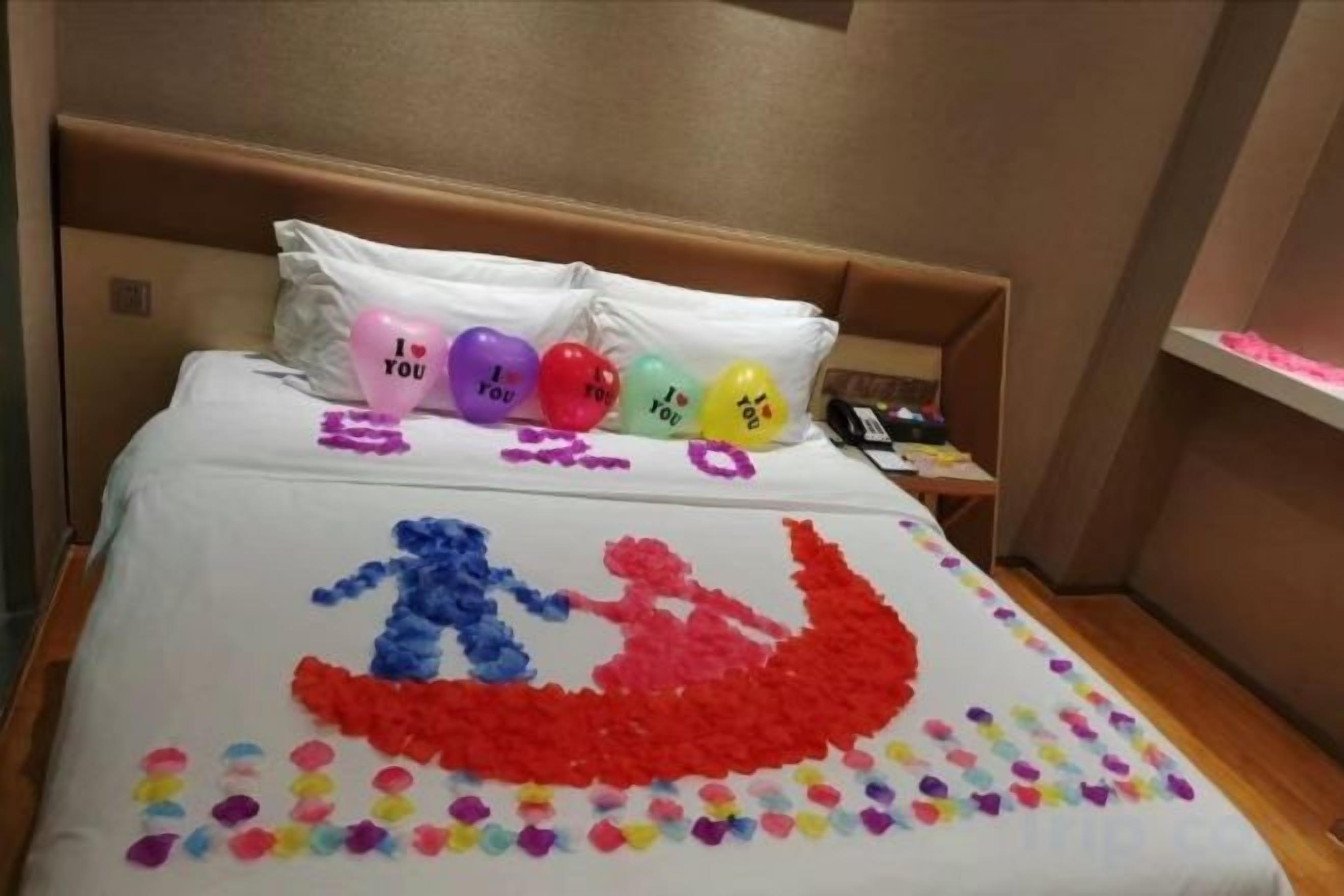 A bed with a white sheet, pillows with heart-shaped decorations, and a bedspread with a boat and people design.
