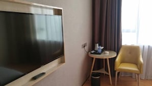 A hotel room with a large flat-screen TV mounted on the wall, a small round table with a chair, a trash bin, and a window with curtains. - Boke Hotel Guangzhou Zhujiang New Town (Guangzhou)