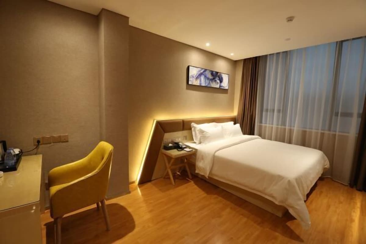 A hotel room with a bed, a desk, a chair, a nightstand, a painting on the wall, and a window with curtains.
