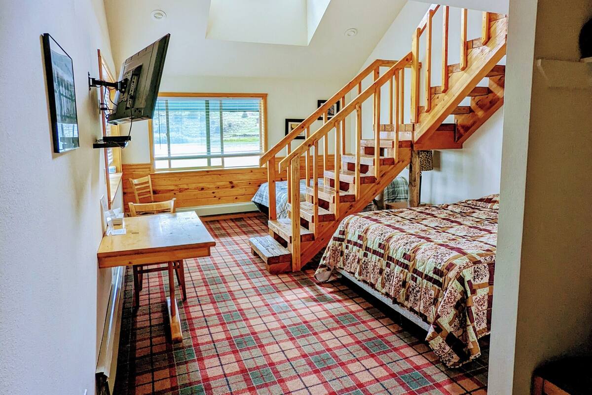 A cozy room with a wooden staircase, a bed with a plaid bedspread, a dining table with chairs, and a TV mounted on the wall.