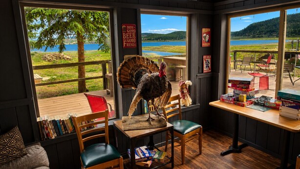 A cozy room with a large window overlooking a lake, a wooden table with board games, a chair with a cushion, and a bookshelf. - Aspen Cove resort (Panguitch)