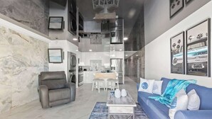 A modern living room with a gray leather chair, a blue sofa, and a coffee table. There is a kitchen area in the background with white cabinets and a dining table. - The Summit 306-Beachfront Getaway on the 3rd Floor (Panama City)