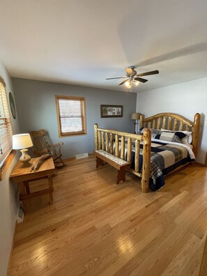 A bedroom with a wooden bed, a bench, a desk with a lamp, a ceiling fan, and a window with blinds. - Waterfront home (Pelican Lake)