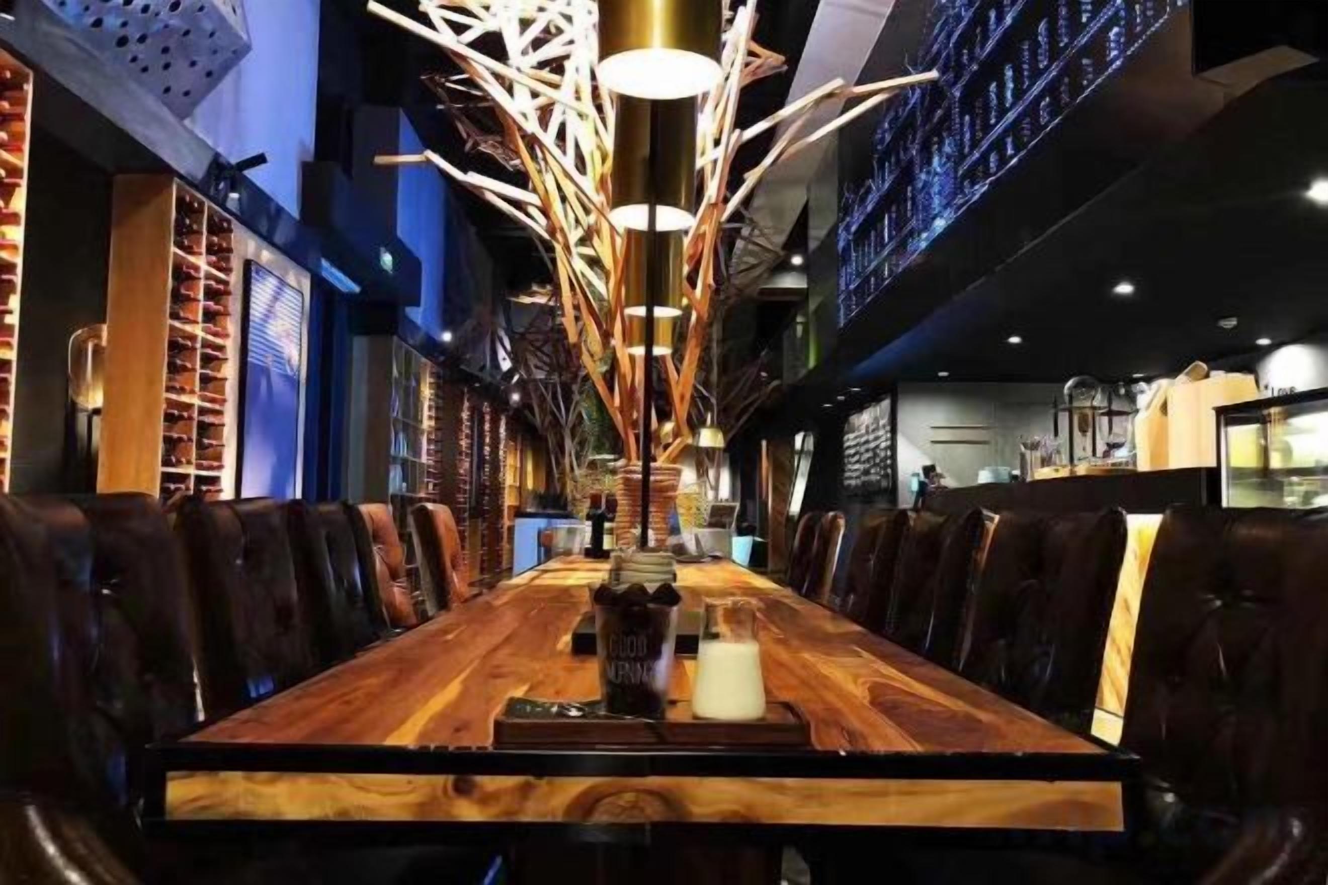 A long wooden dining table with chairs, a modern chandelier, and a bar area in the background.