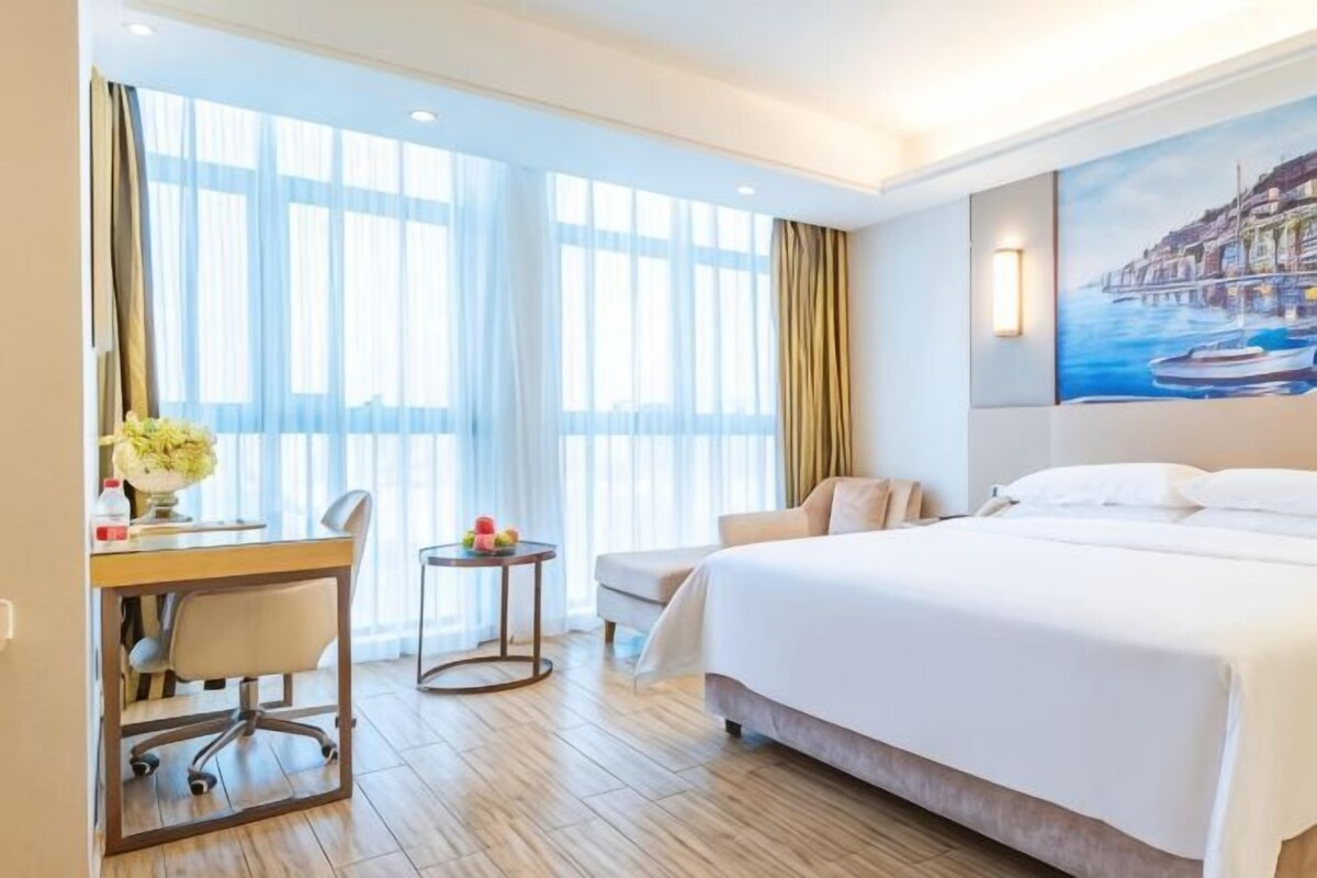 A hotel room with a bed, a desk, a chair, and a small table with a vase of flowers.