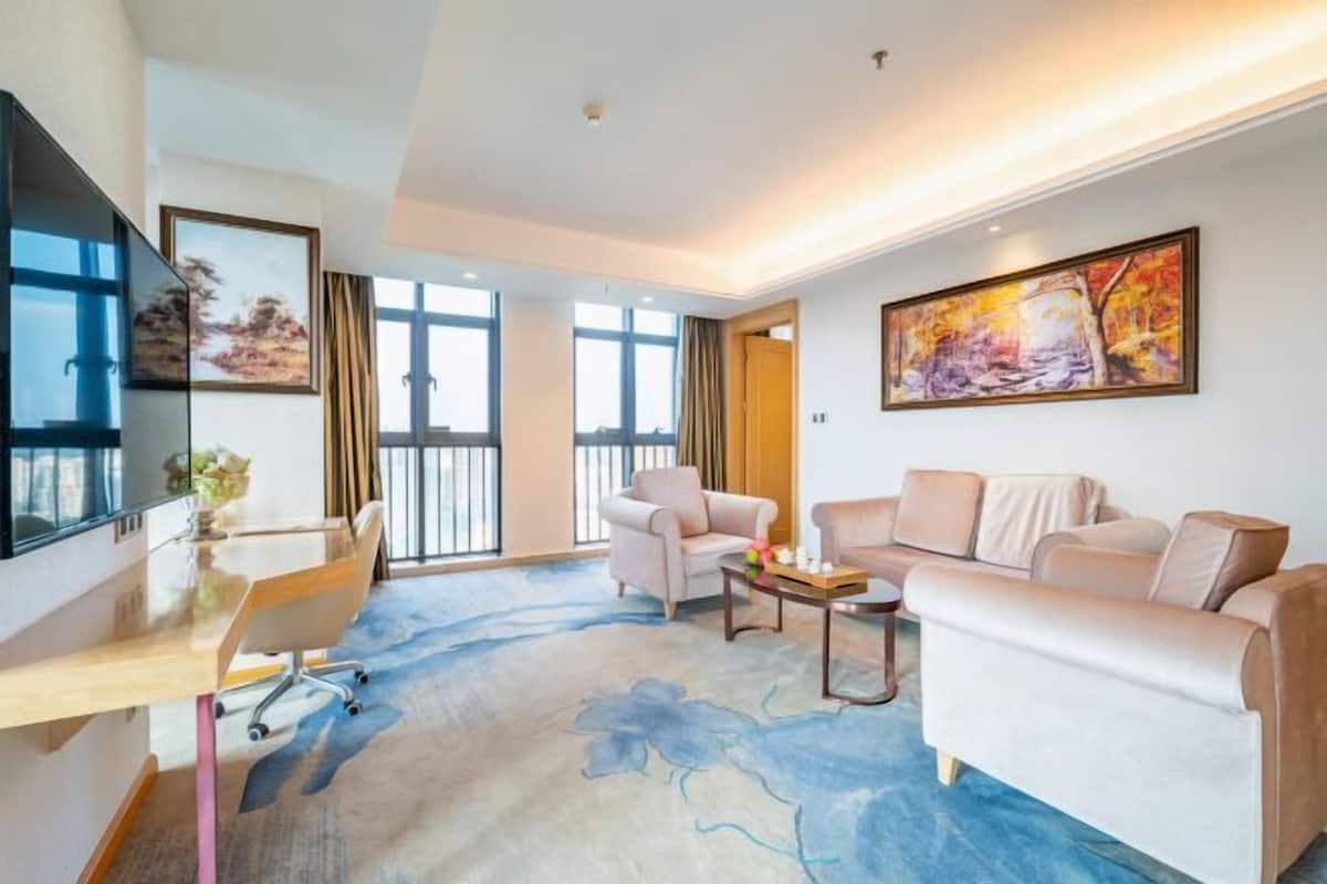A modern hotel room with a large window, a flat-screen TV, a sofa, a chair, a desk, and a painting on the wall.