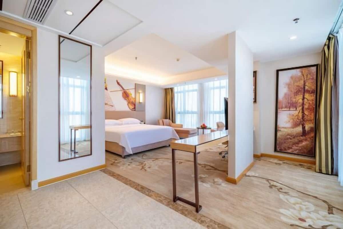 A modern hotel room with a large bed, a sitting area, a dining table, and a view of a park through the window.