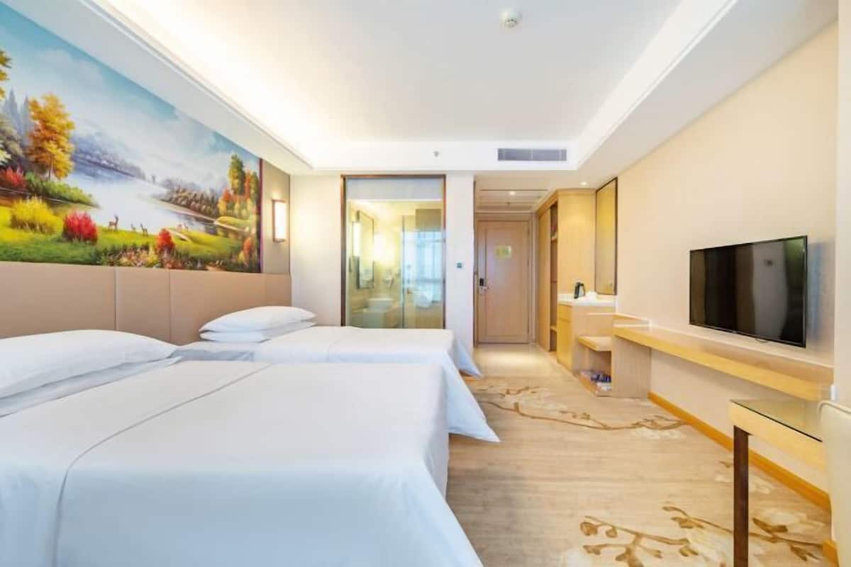 A hotel room with two beds, a TV, a painting on the wall, and a hallway leading to a bathroom.