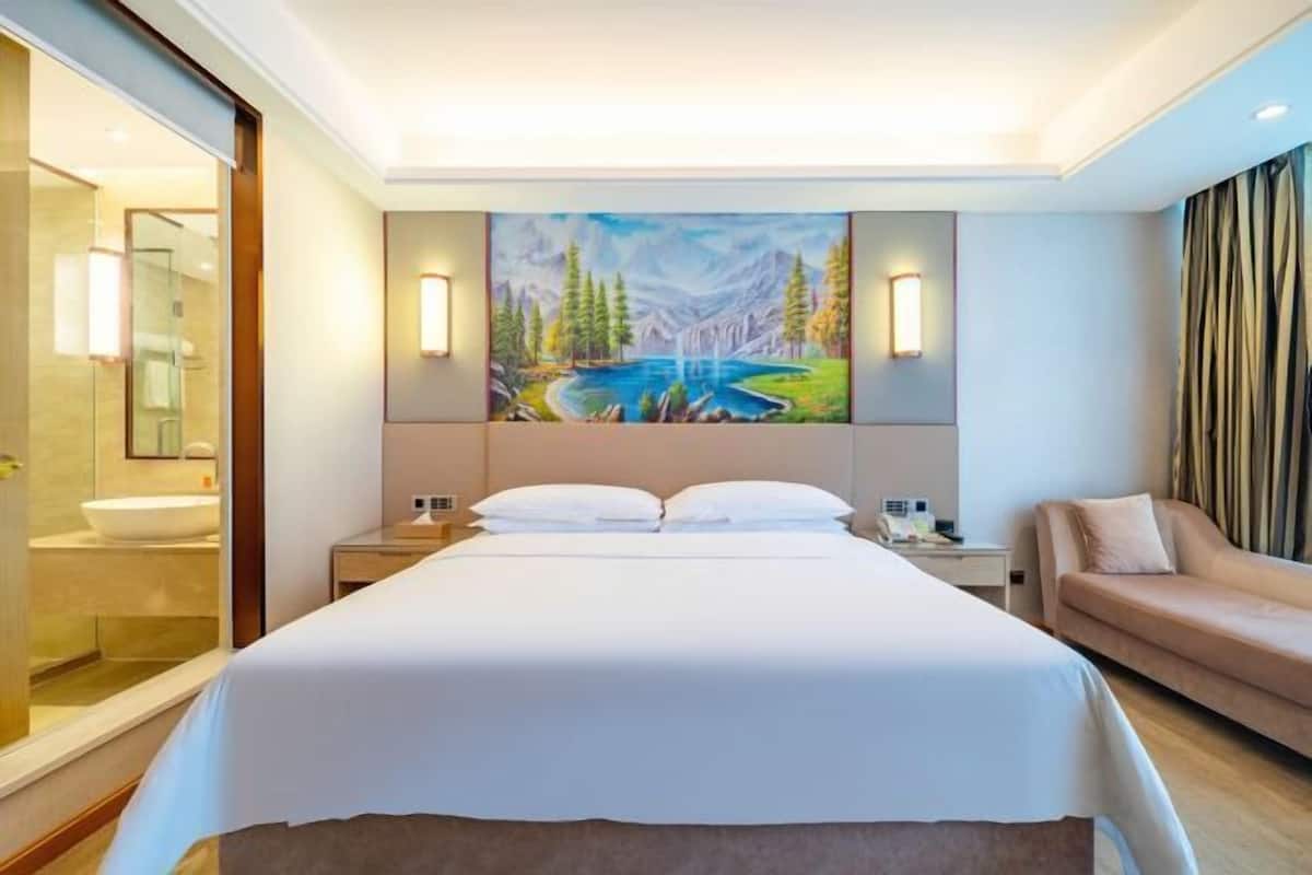 A hotel room with a large bed, a painting of a landscape, a sofa, and a bathroom visible through a glass door.