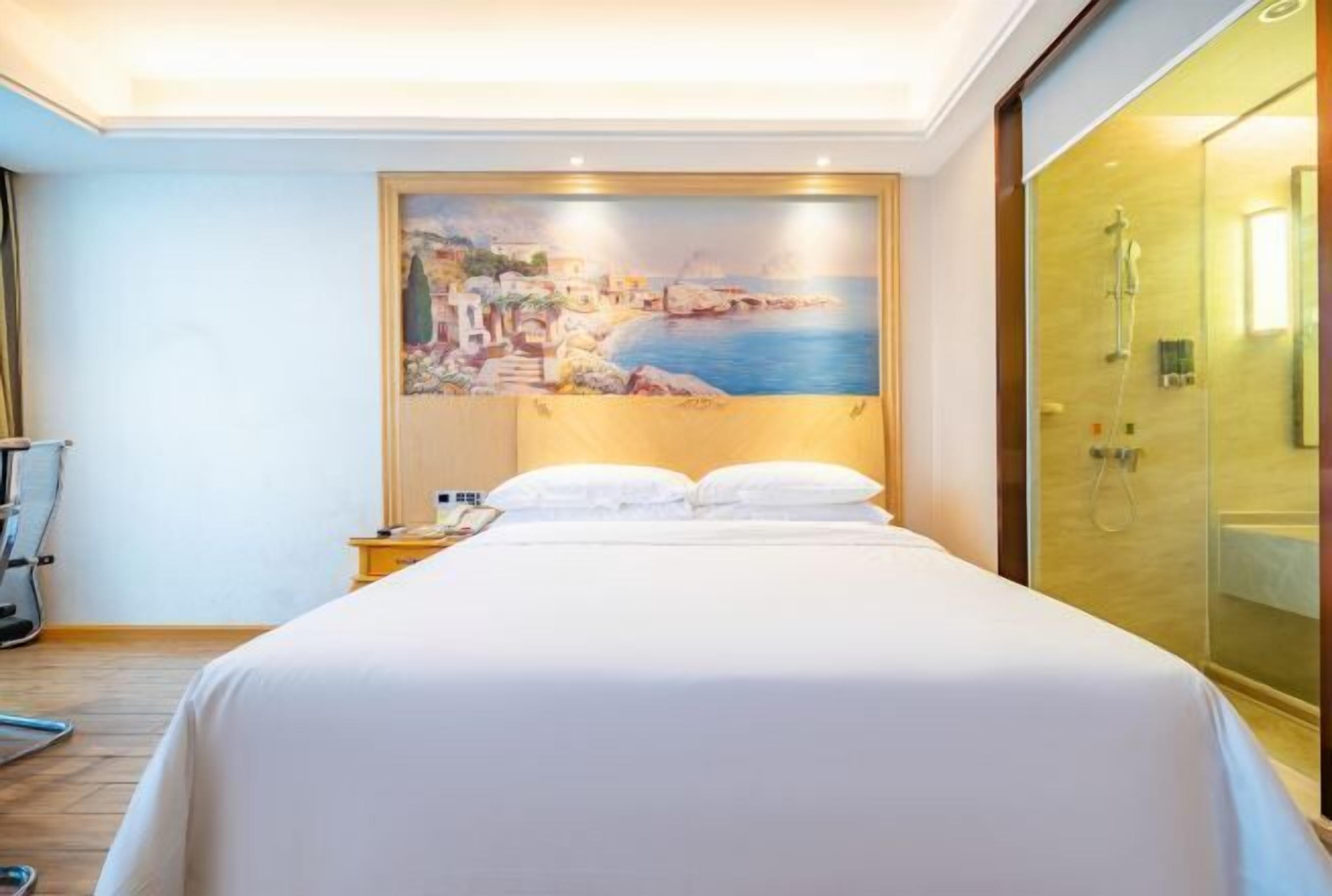 A hotel room with a bed, a painting of a coastal scene, a shower area, and a wooden floor.