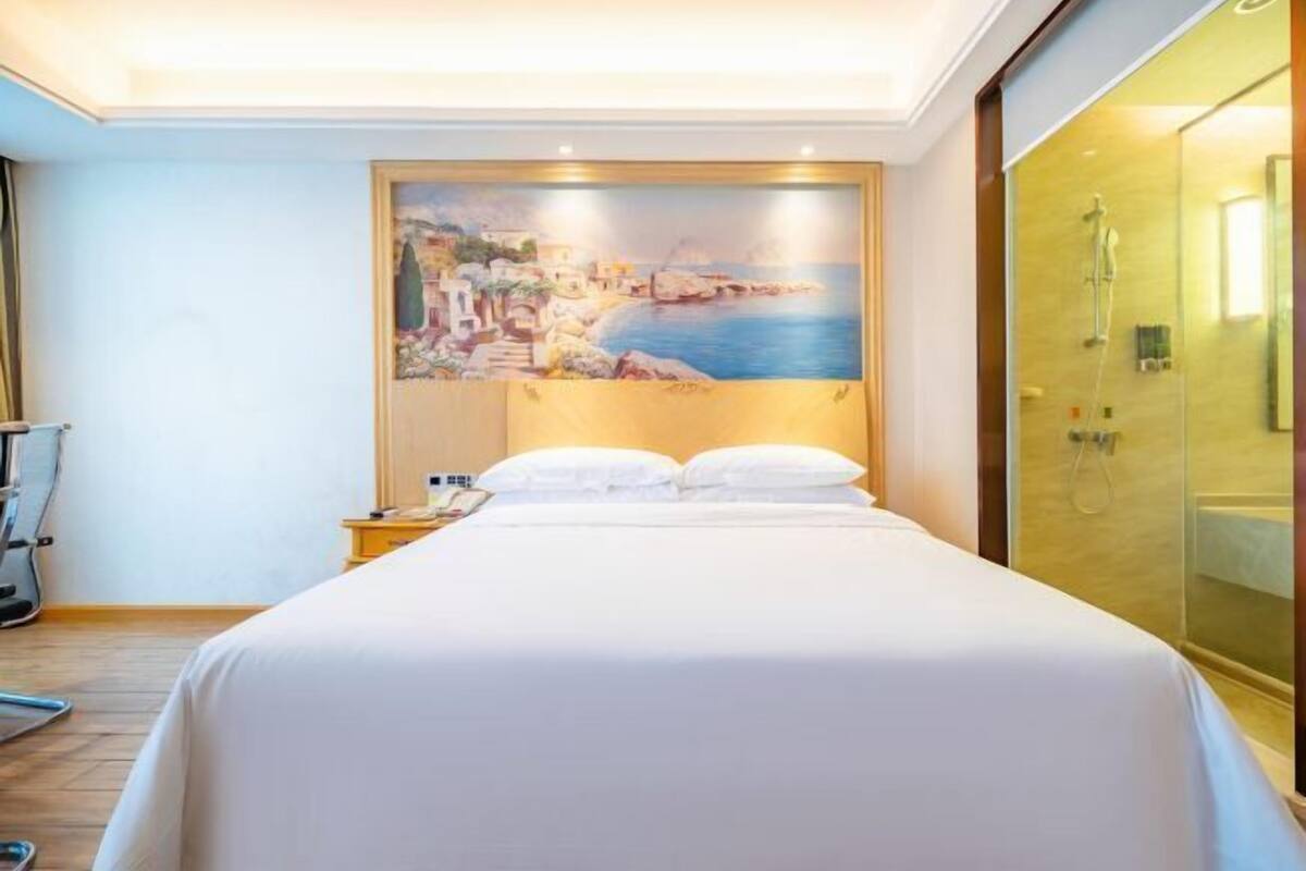 A hotel room with a bed, a painting of a coastal scene, a shower area, and a wooden floor.