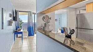 A modern kitchen with a stainless steel refrigerator, a coffee machine, and a sink. There is a blue table with a chair and a blue curtain in the background. - Sunbird 811E - Beautiful Beachfront Getaway! (Panama City Beach)