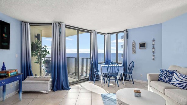 A room with a balcony, a dining table with chairs, a sofa, and a TV mounted on the wall. - Sunbird 811E - Beautiful Beachfront Getaway! (Panama City Beach)