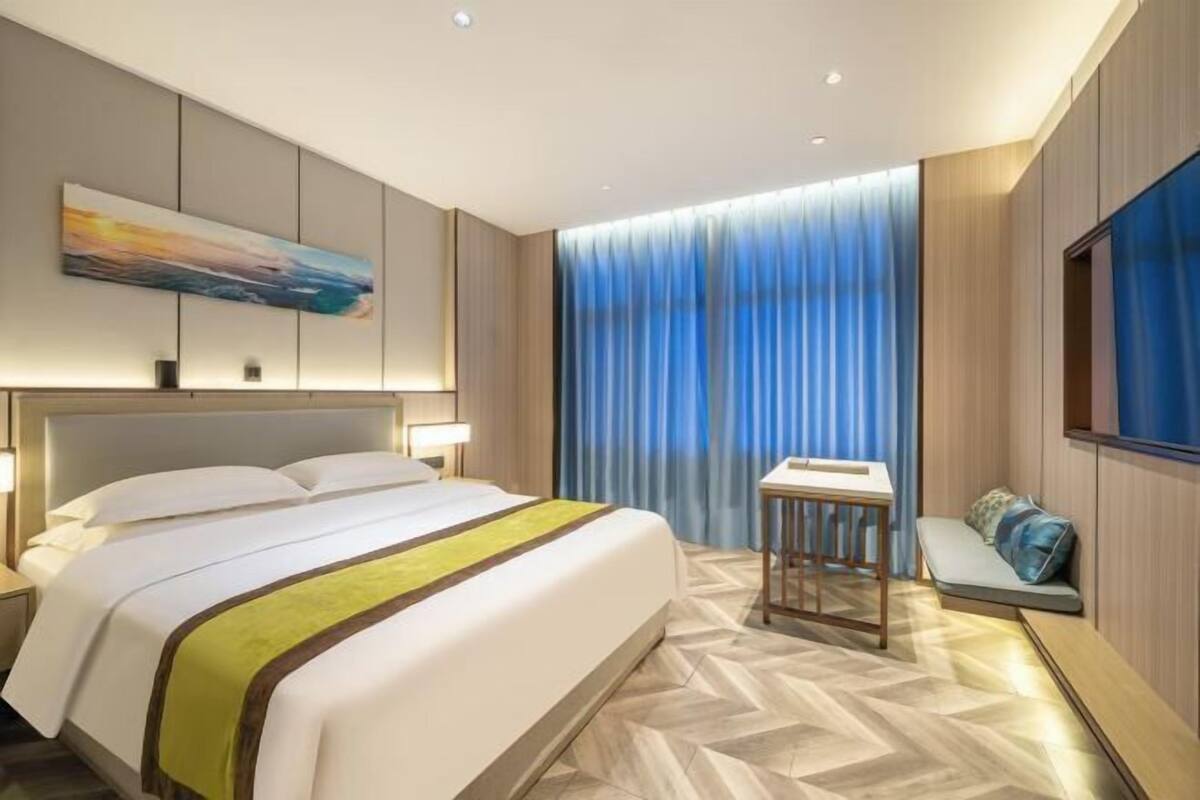 Una habitación de hotel moderna con una cama grande, una mesita de noche, una pequeña zona de estar y un amplio ventanal con cortinas.