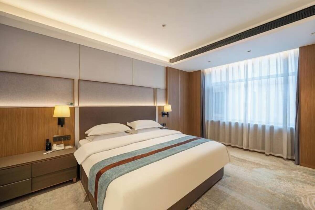 Una habitación de hotel con una cama grande, lámparas de noche, una mesita de noche y un ventanal con cortinas.