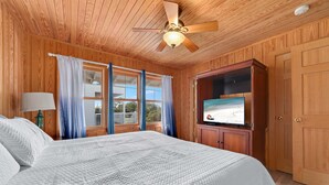 Ein Schlafzimmer mit einem großen Bett, Holzwänden, einem Deckenventilator, einem Fernseher und einem Fenster mit Vorhängen.