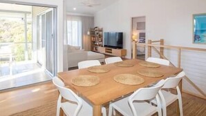 A modern dining area with a wooden table and white chairs, a television, and a bookshelf in the background. - The Lookout Beach House - Gorgeous renovated 2 level home with spectacular ocean views (Point Lookout)
