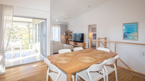 A modern living room with a wooden dining table, white chairs, a flat-screen TV, and a bookshelf. - The Lookout Beach House - Gorgeous renovated 2 level home with spectacular ocean views (Point Lookout)