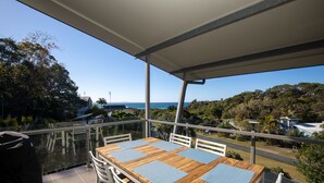 A wooden table with blue placemats, white chairs, and a glass balustrade overlooking a scenic view of the ocean and greenery. - The Lookout Beach House - Gorgeous renovated 2 level home with spectacular ocean views (Point Lookout)
