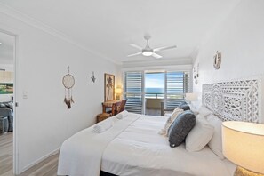 A bedroom with a bed, a ceiling fan, a nightstand, a lamp, and a view of the ocean through the window.