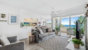 A modern living room with a grey sofa, a small round table, and a large window overlooking the ocean. - The Cove - Jewel by the Sea (Point Lookout)